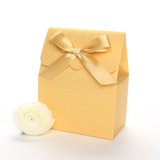 Lux Party’s gold favor box with a scalloped edge and a gold satin bow next to white ranunculus flowers.