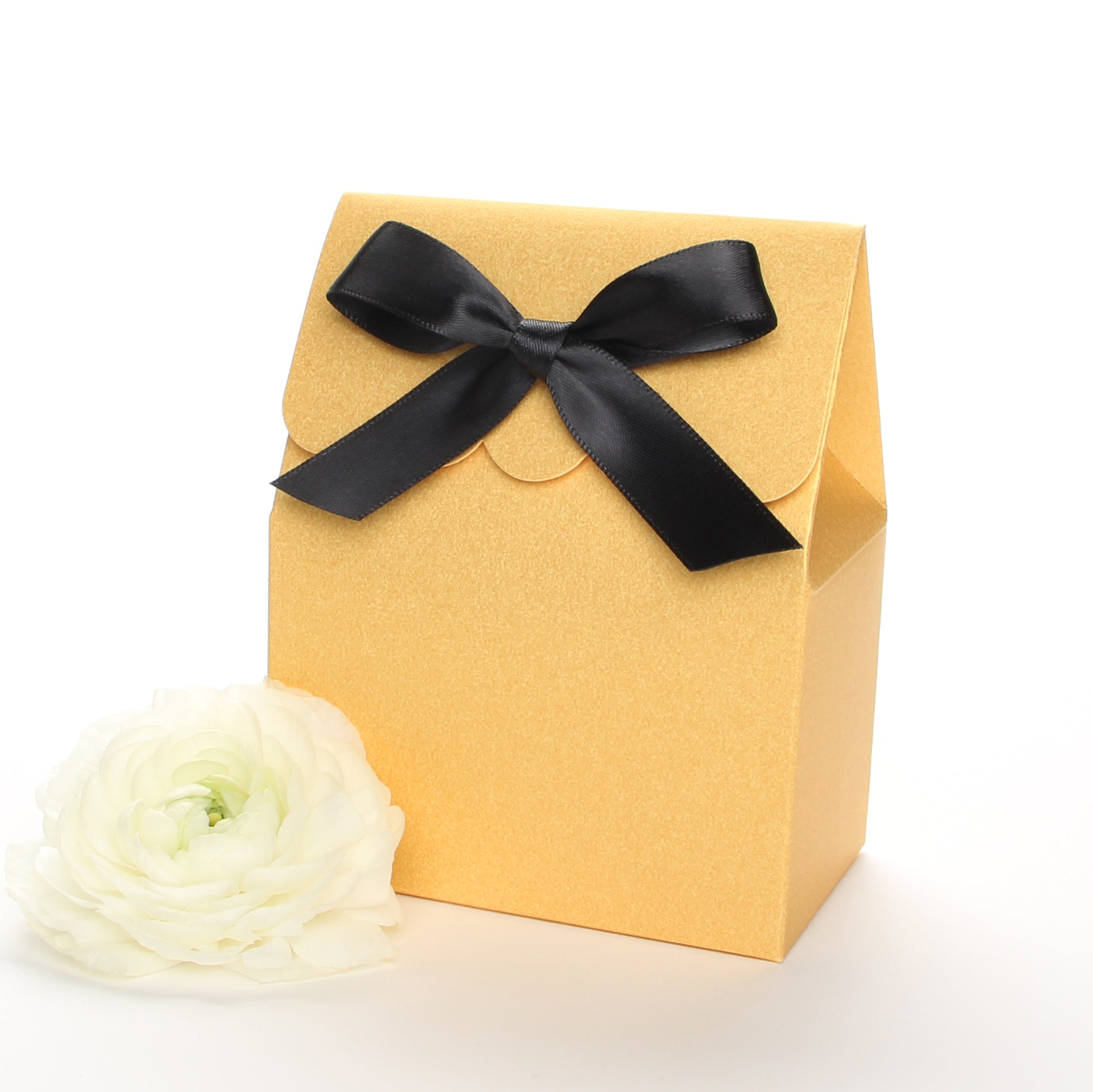 Lux Party’s gold favor box with a scalloped edge and a black satin bow next to white ranunculus flowers.