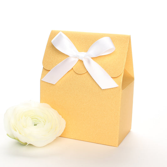 Lux Party’s gold favor box with a scalloped edge and a white satin bow next to white ranunculus flowers.