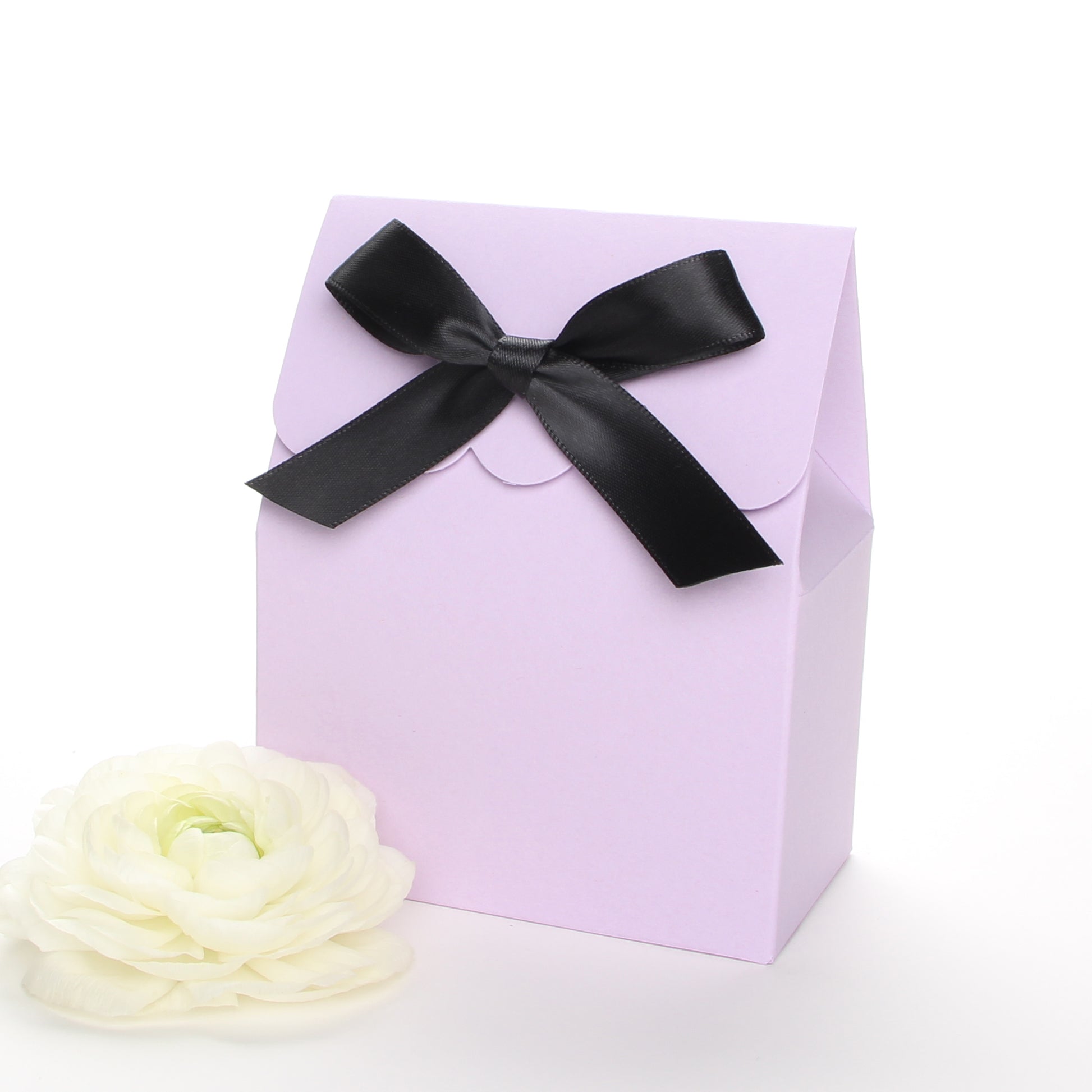 Lux Party’s lavender favor box with a scalloped edge and a black satin bow next to pink and white ranunculus flowers.