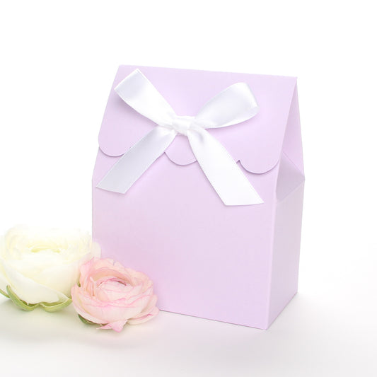 Lux Party’s lavender favor box with a scalloped edge and a white satin bow next to pink and white ranunculus flowers.