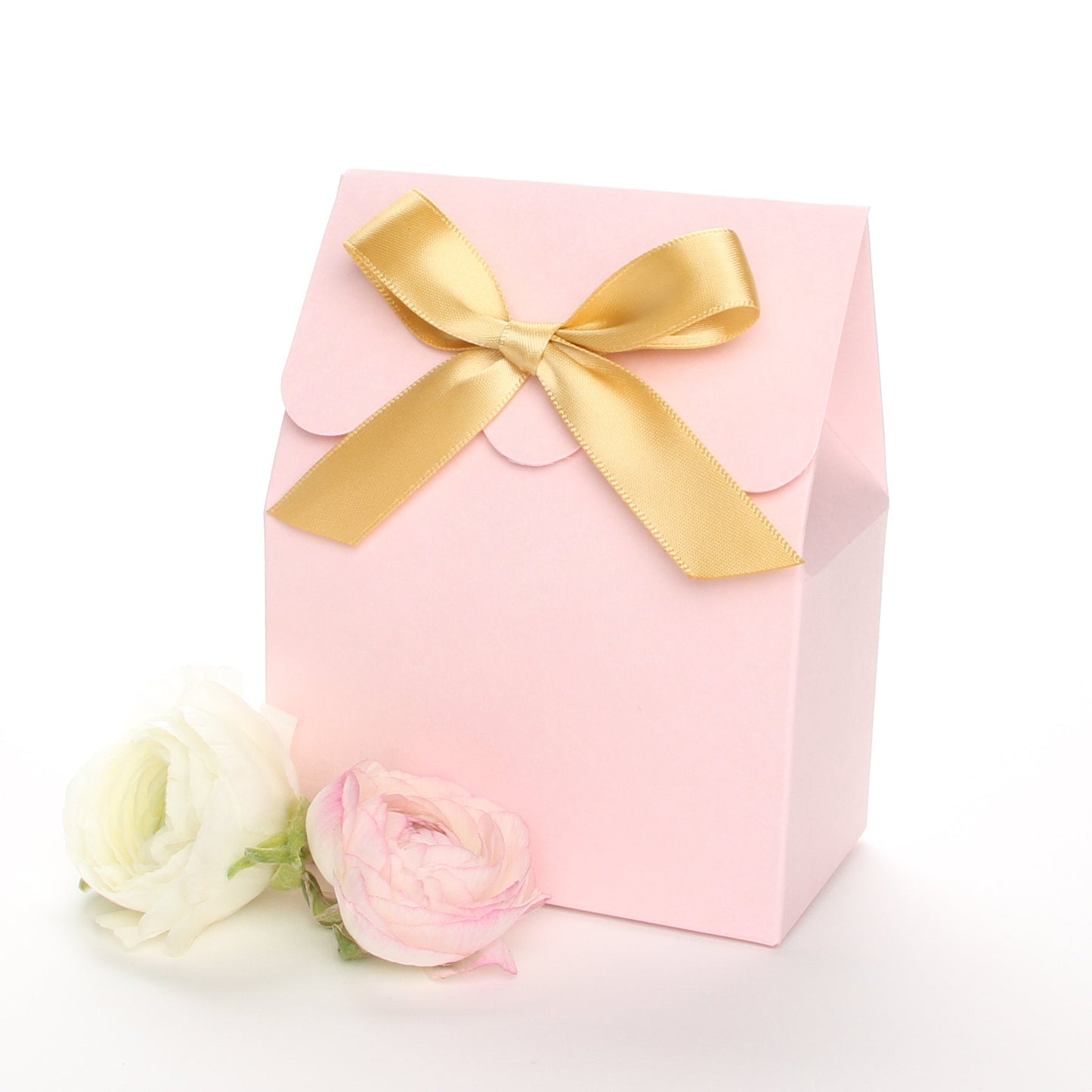 Lux Party’s pink favor box with a scalloped edge and a gold bow next to pink and white ranunculus flowers.