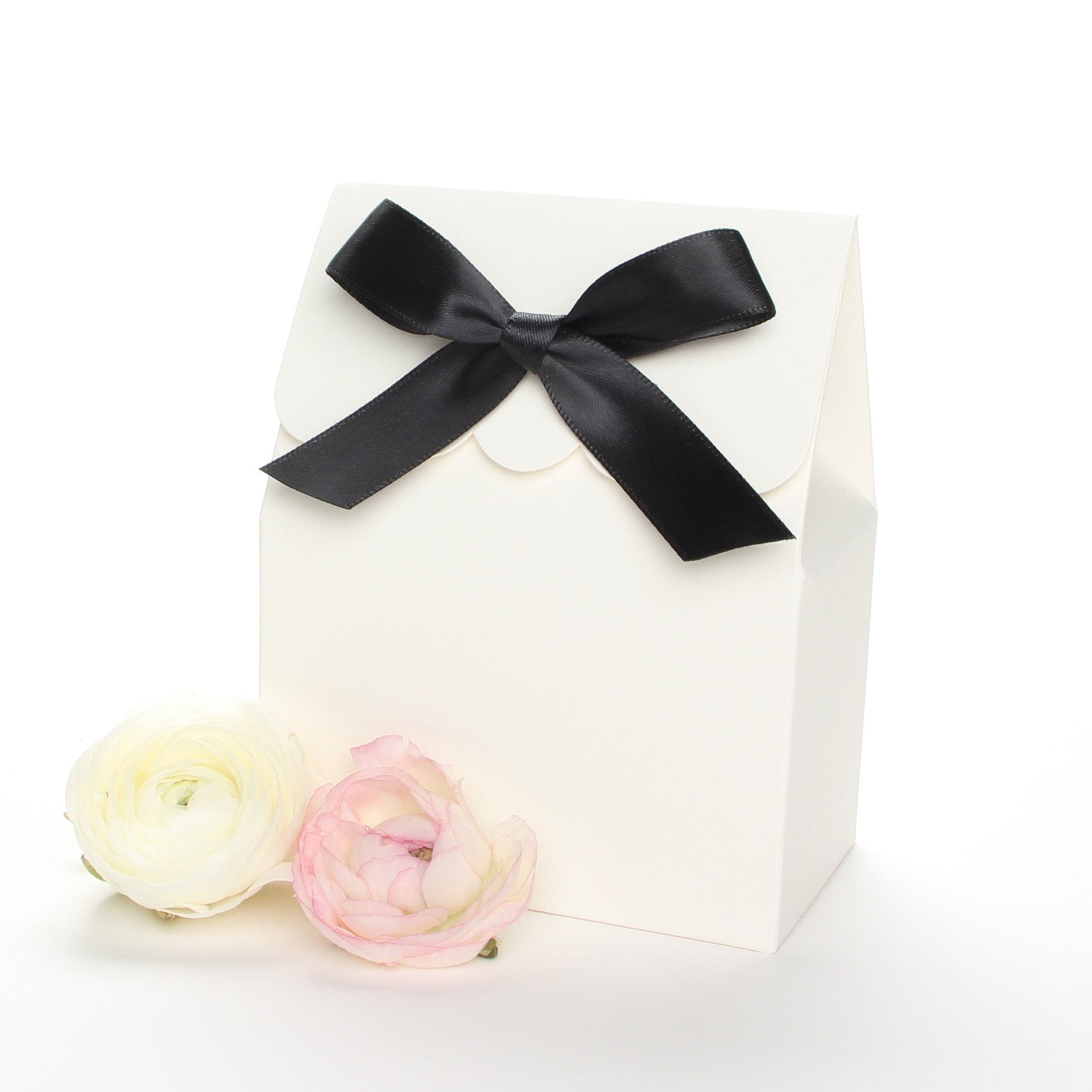 Lux Party’s ivory favor box with a scalloped edge and a black satin bow next to pink and white ranunculus flowers.