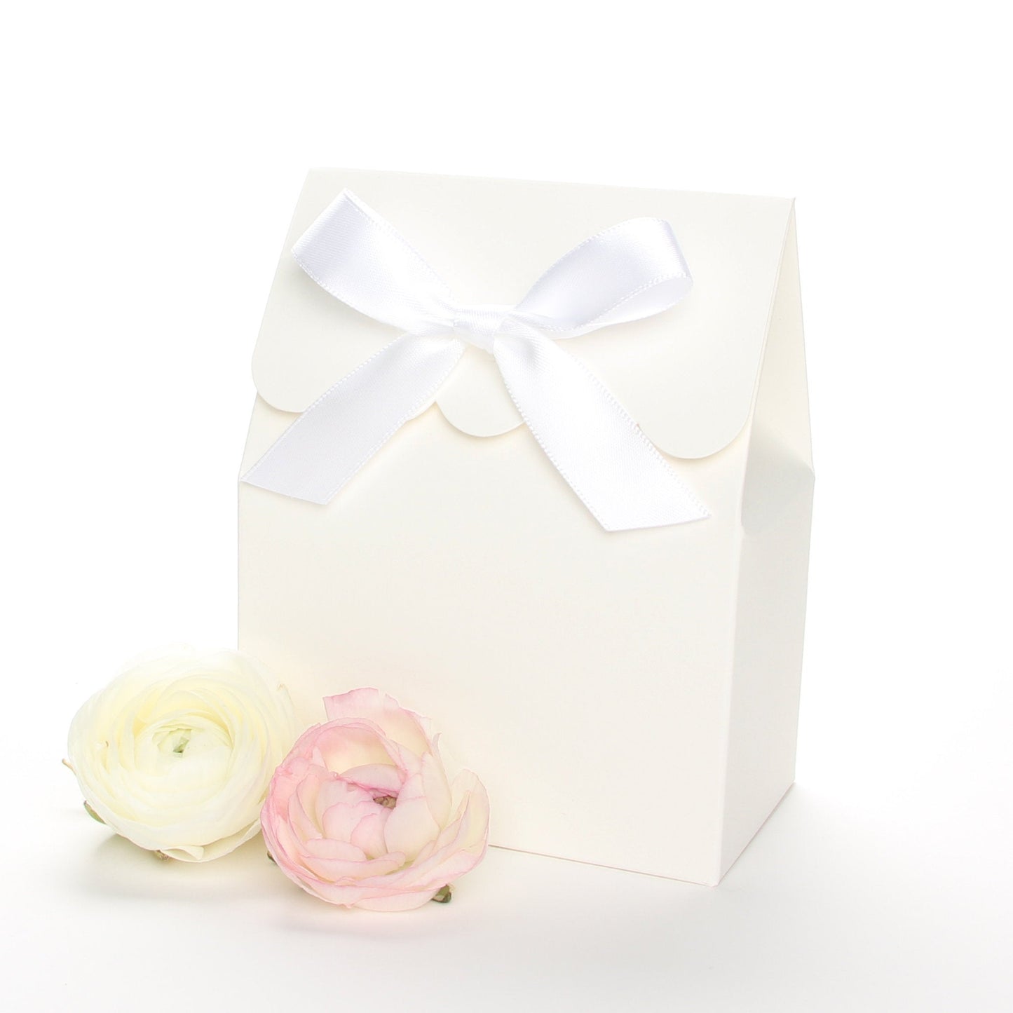 Lux Party’s ivory favor box with a scalloped edge and a white satin bow next to pink and white ranunculus flowers.