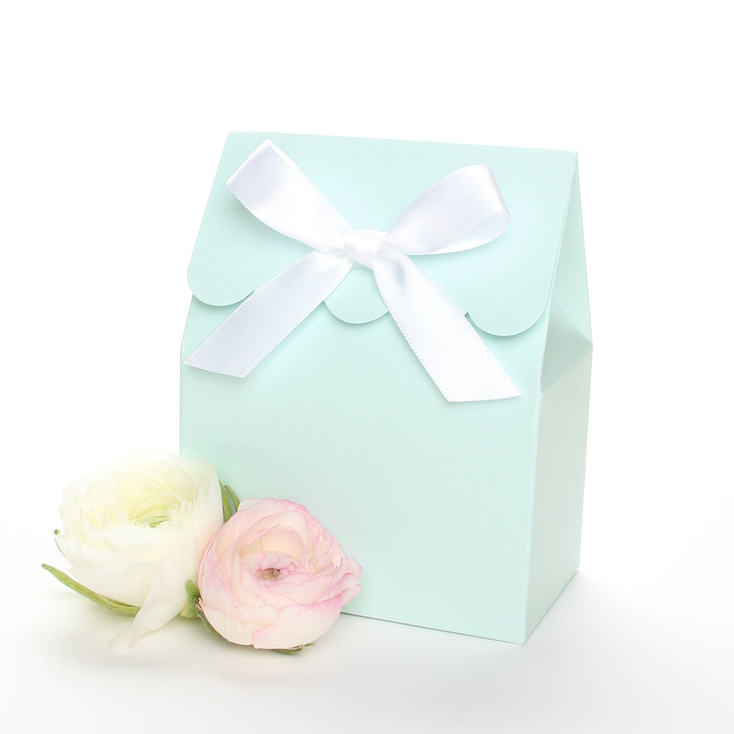 Lux Party’s light blue favor box with a scalloped edge and a white satin bow next to pink and white ranunculus flowers.