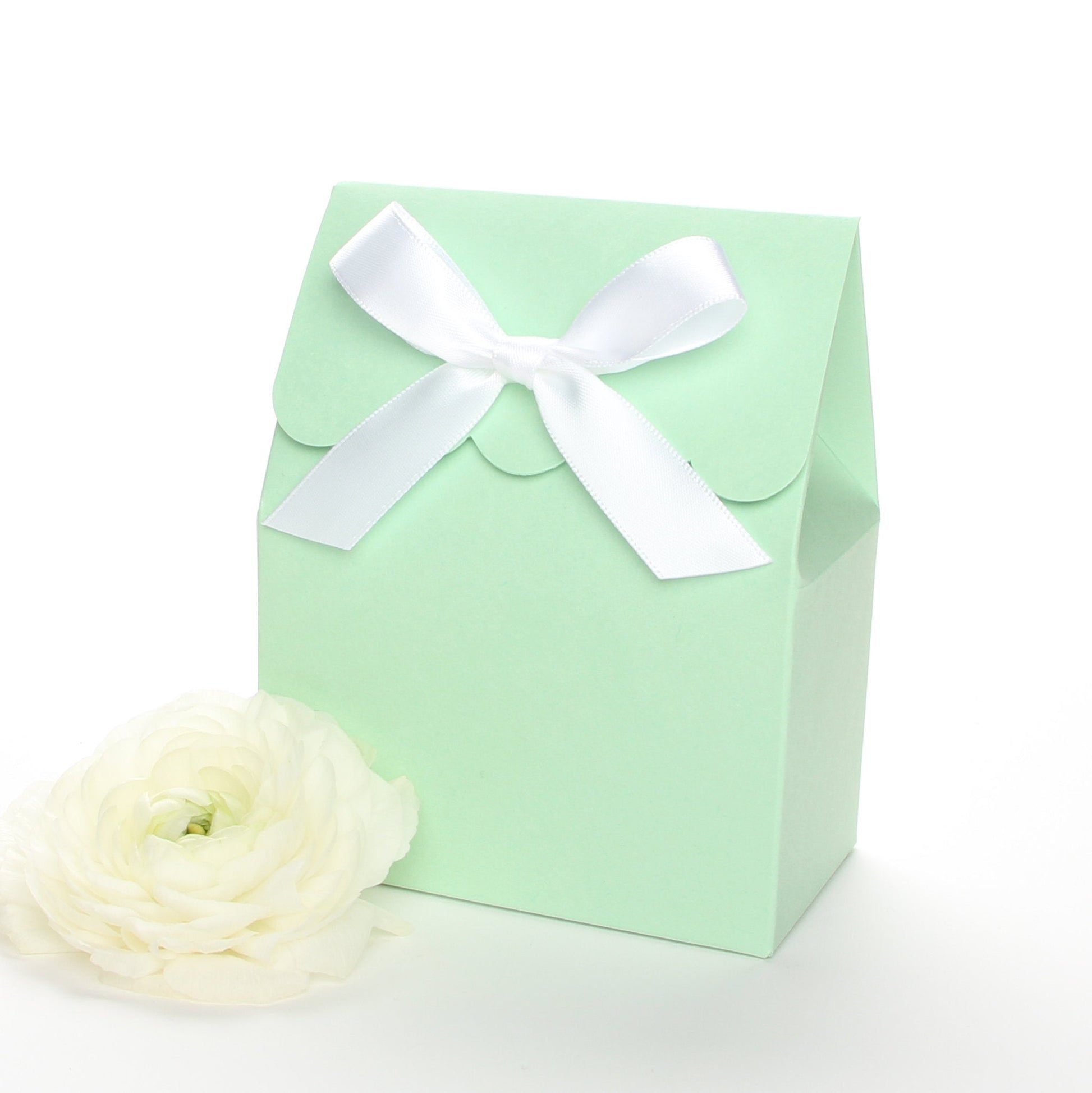 Lux Party’s mint green favor box with a scalloped edge and a white satin bow next to white ranunculus flowers.