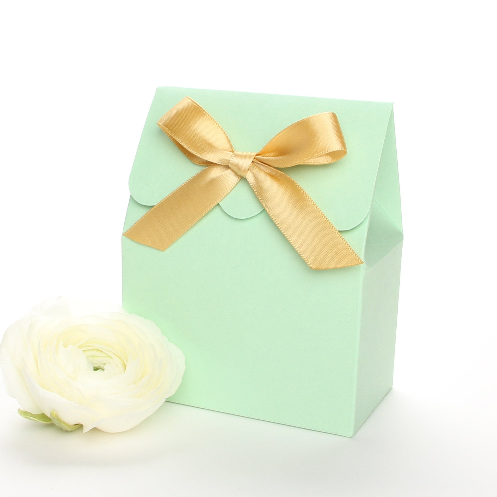 Lux Party’s mint green favor box with a scalloped edge and a gold satin bow next to white ranunculus flowers.
