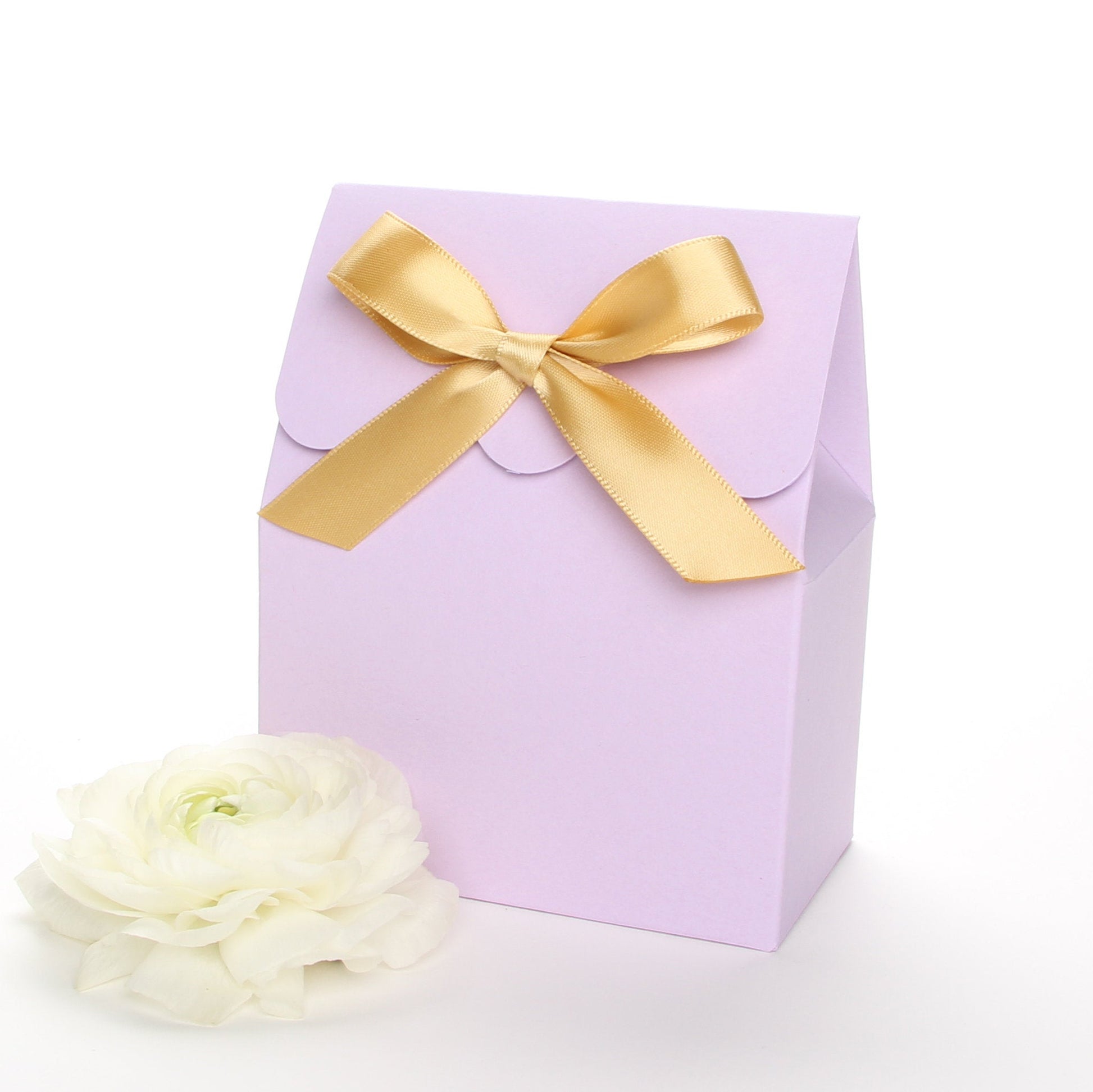 Lux Party’s lavender favor box with a scalloped edge and a gold satin bow next to white ranunculus flowers.