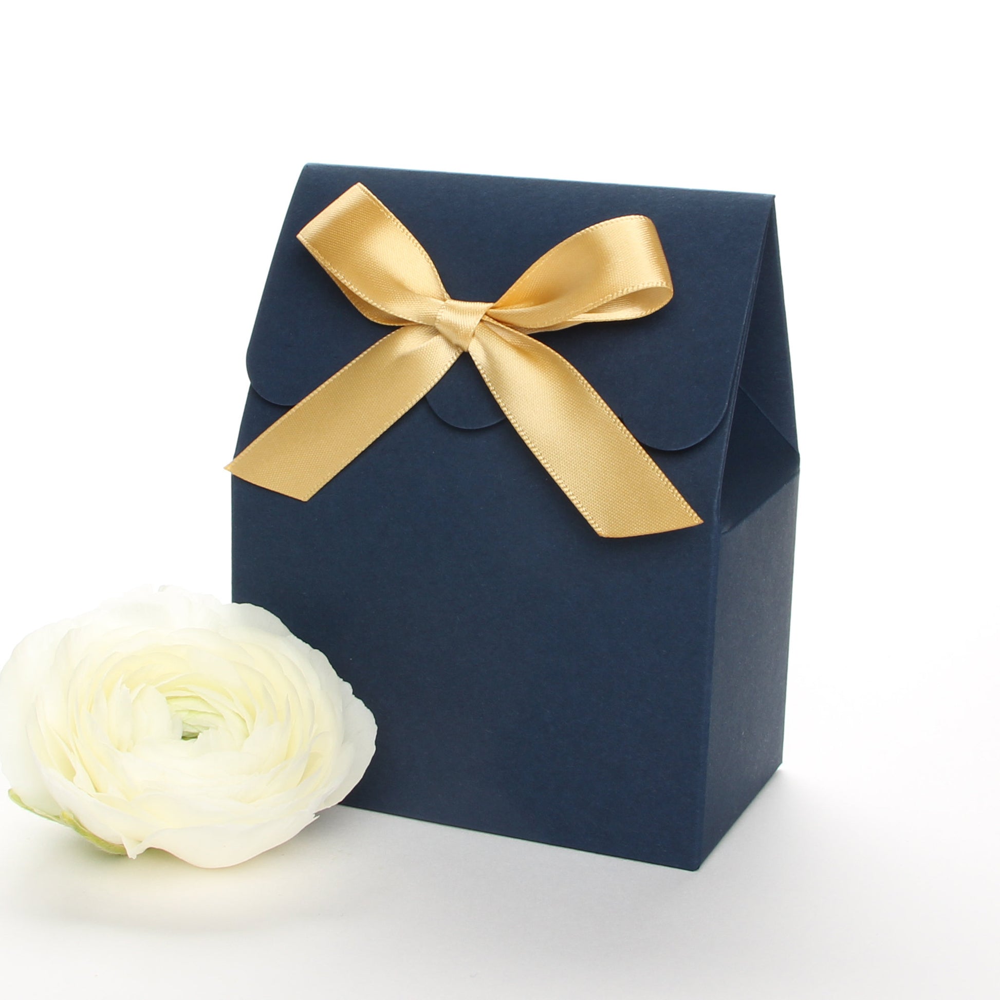 Lux Party’s navy blue favor box with a scalloped edge and a gold bow next to white ranunculus flowers.