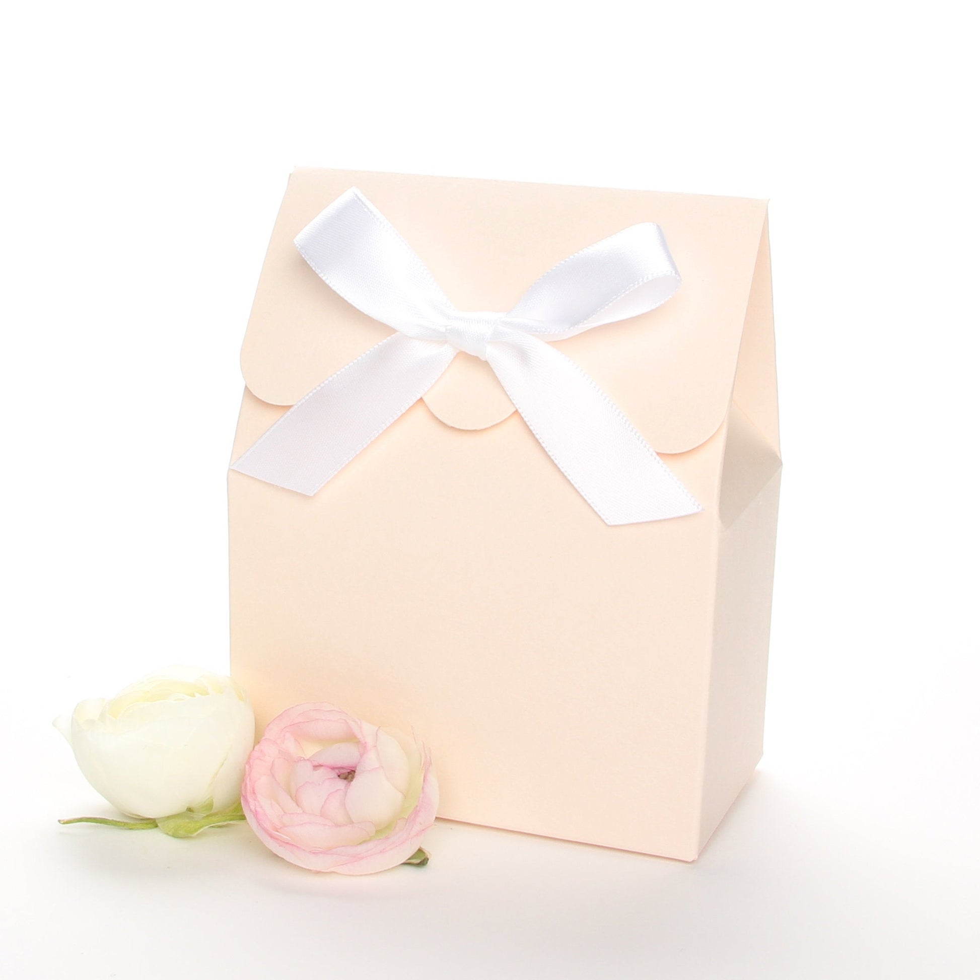 Lux Party’s blush favor box with a scalloped edge and a white bow next to pink and white ranunculus flowers.