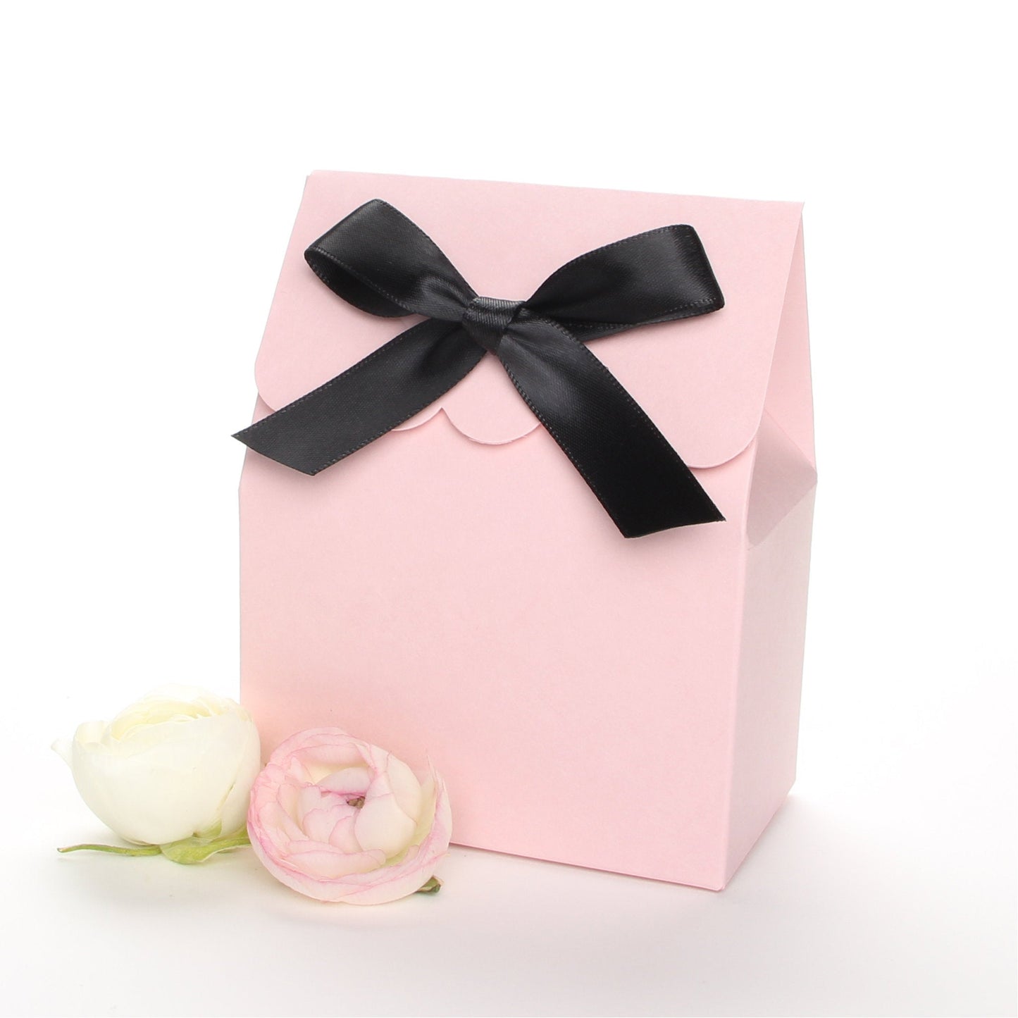 Lux Party’s pink favor box with a scalloped edge and a black satin bow next to pink and white ranunculus flowers.