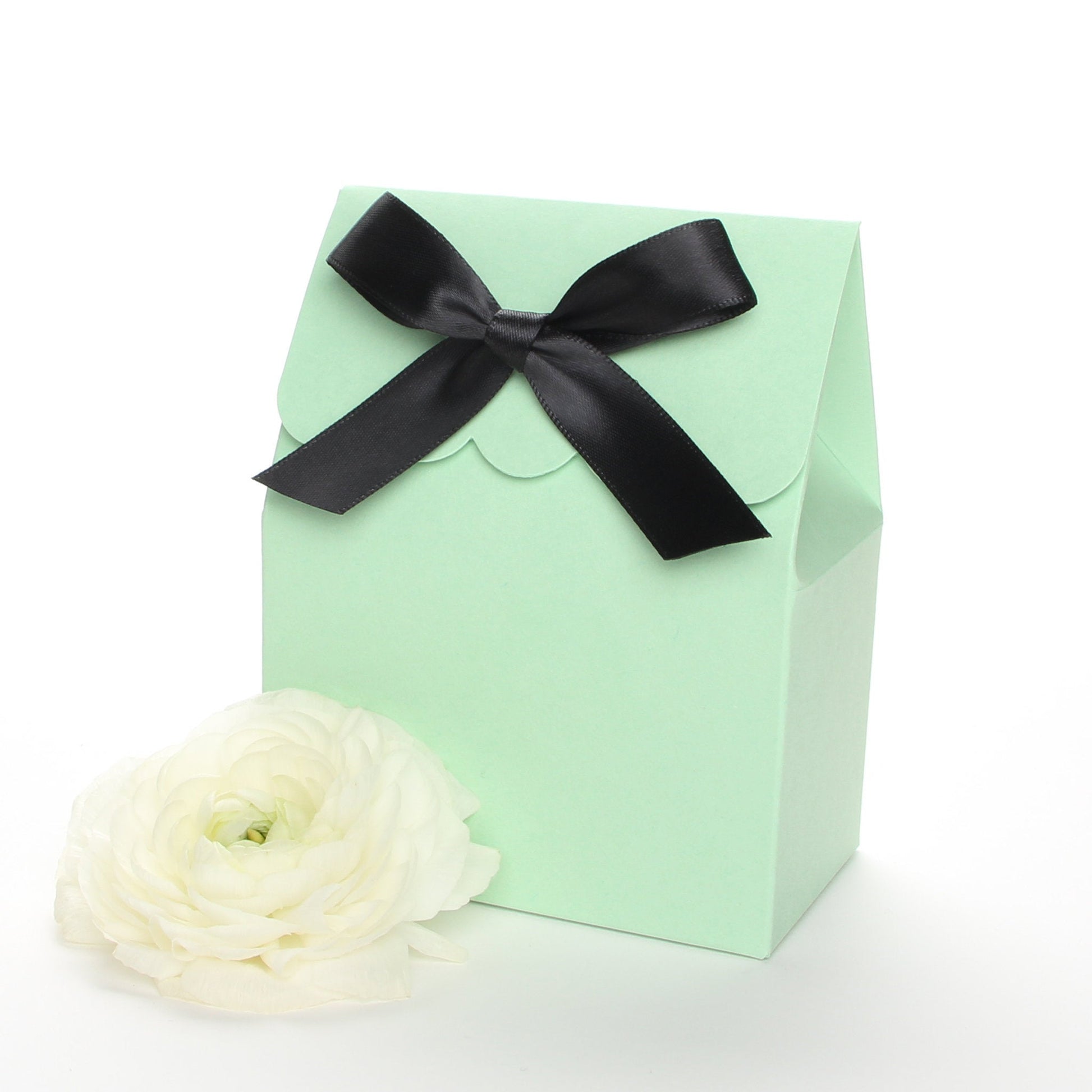 Lux Party’s mint green favor box with a scalloped edge and a black satin bow next to white ranunculus flowers.