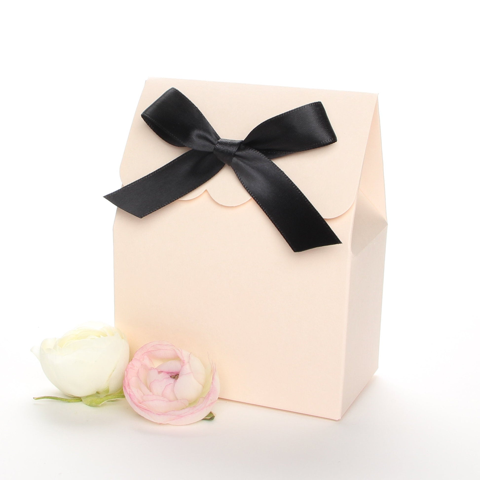 Lux Party’s blush favor box with a scalloped edge and a black bow next to white ranunculus flowers.