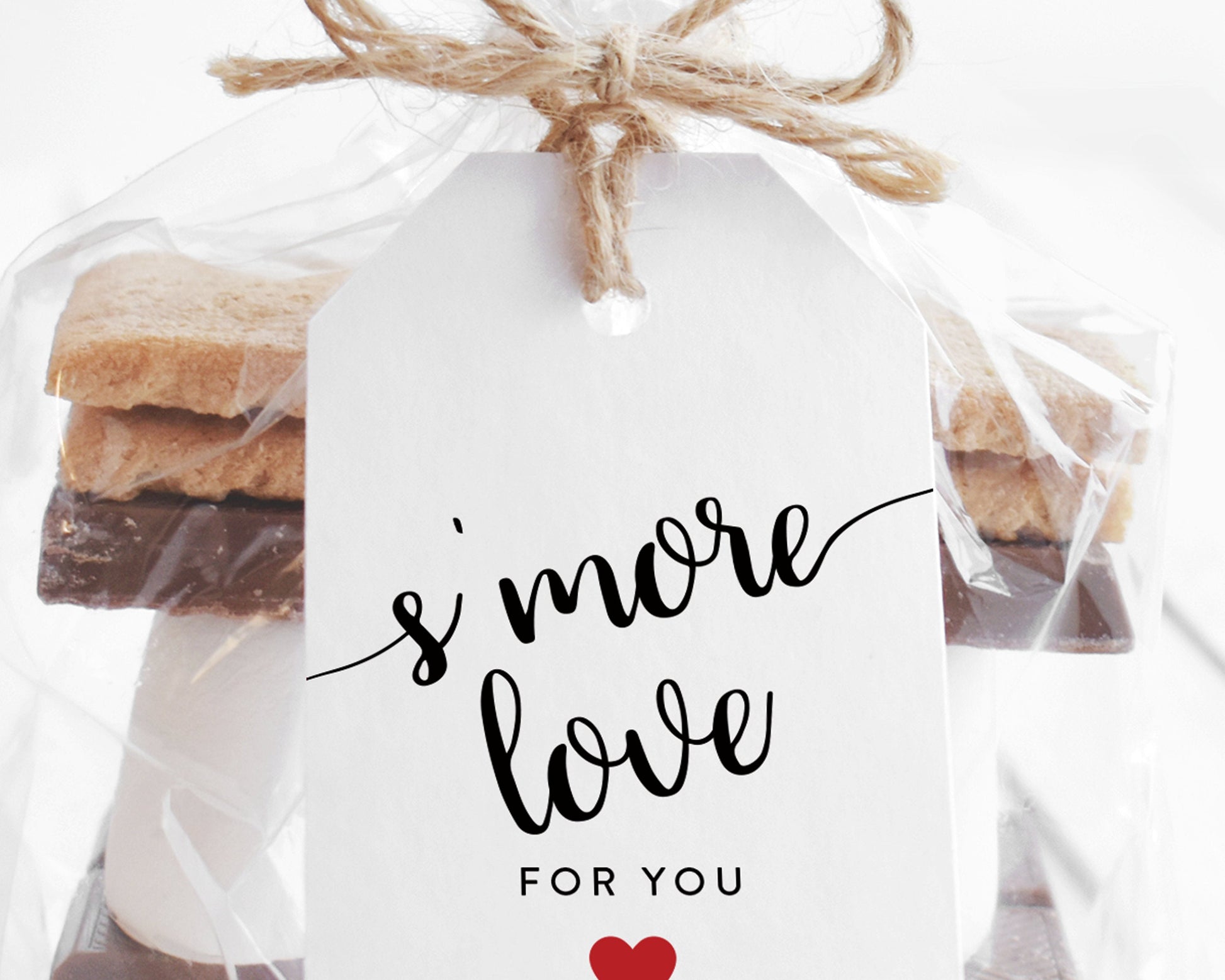 Close up of Lux Party’s white s’more love wedding favor tag with black script and a red heart, tied to a clear favor bag of s’mores ingredients.